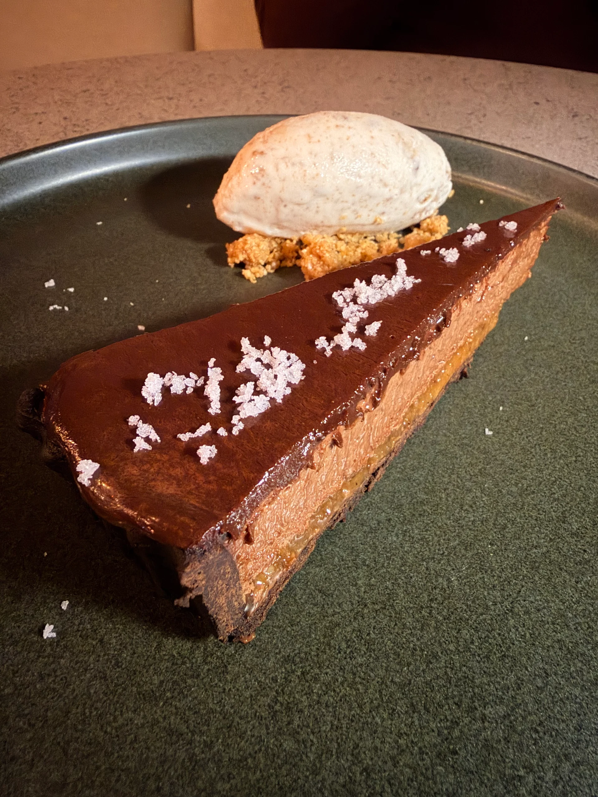 A decadent slice of chocolate cheesecake topped with a scoop of vanilla ice cream and sprinkled with sea salt, presented on a dark green plate.