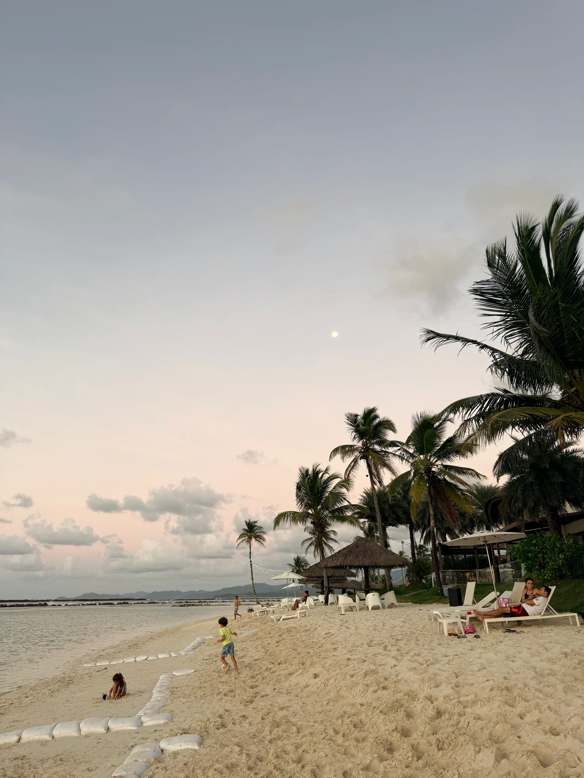 Une scène sereine du soir sur la plage de Playa Escondida, à Panama. Les palmiers se balancent doucement sous un ciel crépusculaire, avec une pleine lune visible au-dessus. Les familles profitent de la plage, certaines construisant des châteaux de sable, tandis que d'autres se détendent dans des chaises longues près de paillotes aux toits de paille. Le sable doux et l'eau calme créent une atmosphère tropicale et paisible.