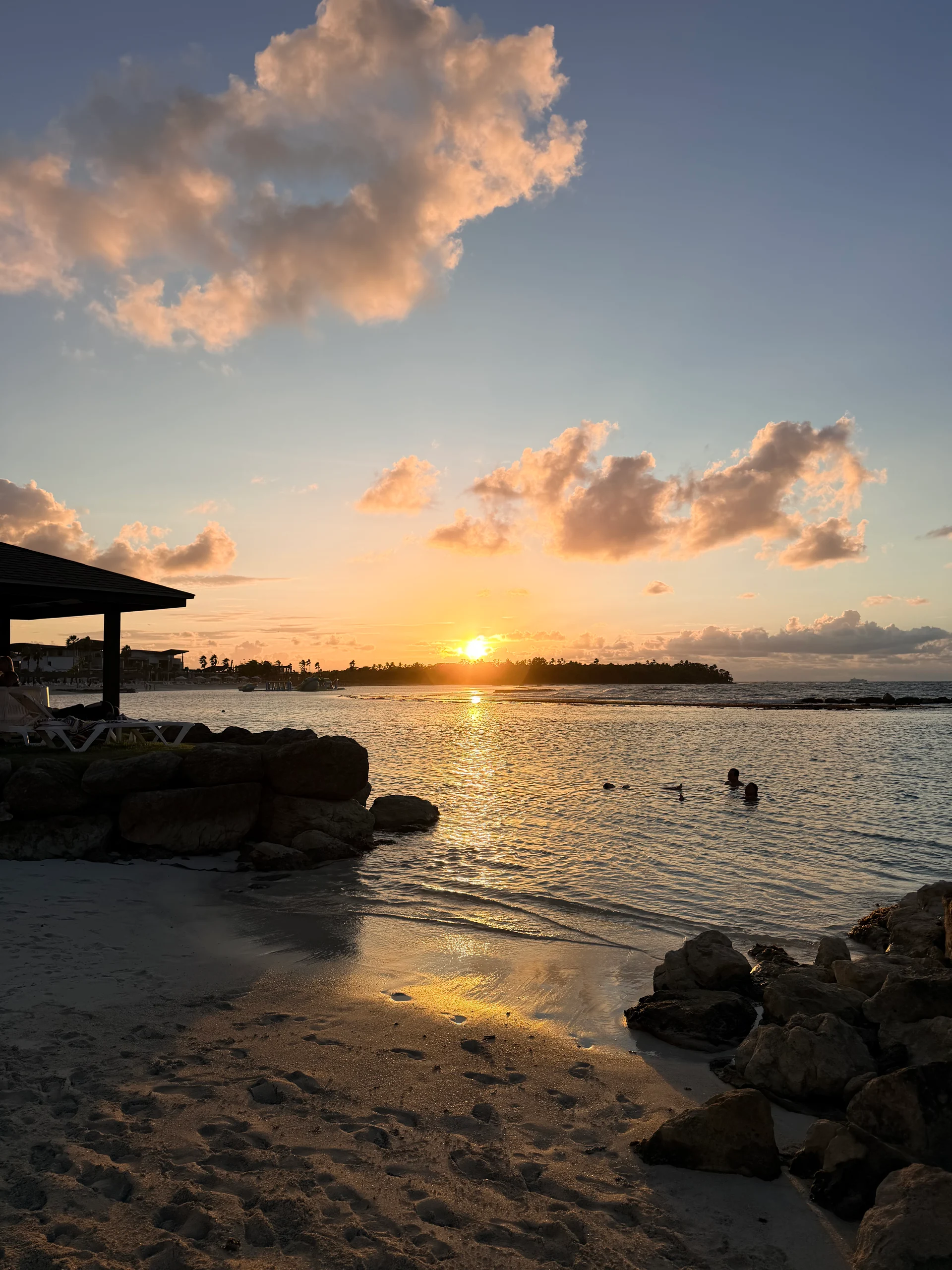 Une vue imprenable sur le coucher de soleil au-dessus de la mer des Caraïbes à Playa Escondida, Panama. La lumière dorée se reflète sur les eaux calmes, mettant en valeur le rivage rocheux et la silhouette des palmiers et des bâtiments au loin. Plusieurs personnes nagent dans l'océan, profitant de l'atmosphère paisible.