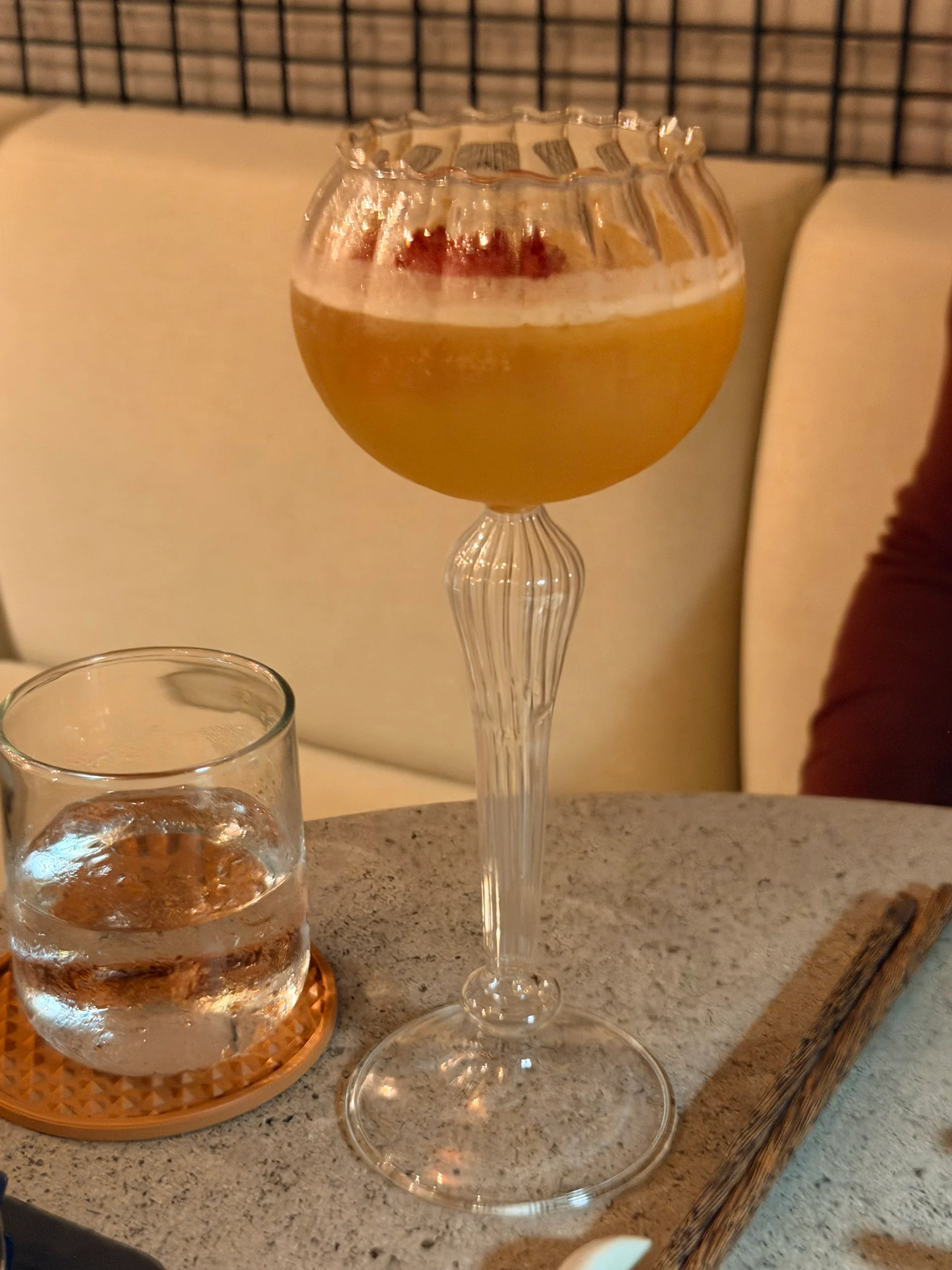 A vibrant orange cocktail with a foamy top sits in an elegant, ridged coupe glass, accompanied by a glass of water with ice. The drink is presented on a textured gray surface with a glimpse of a wooden stick and a blurred background suggesting a modern restaurant setting.