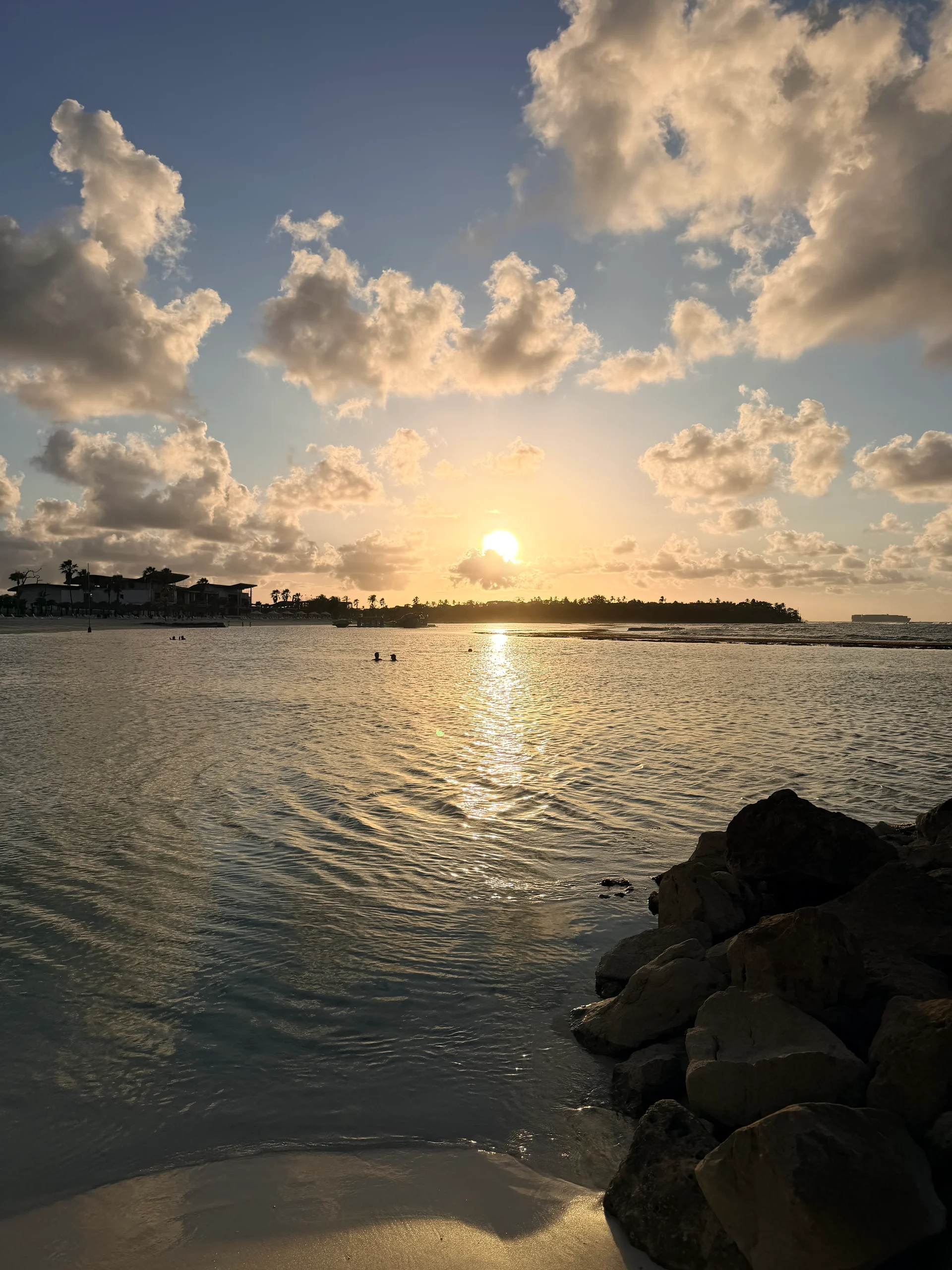 Une vue imprenable sur le coucher de soleil au-dessus des eaux turquoise calmes de Playa Escondida, Panama. Le soleil doré disparaît sous l'horizon, se reflétant brillamment sur la surface de l'eau, encadré par une ligne de rochers usés au premier plan et un resort immaculé au loin.