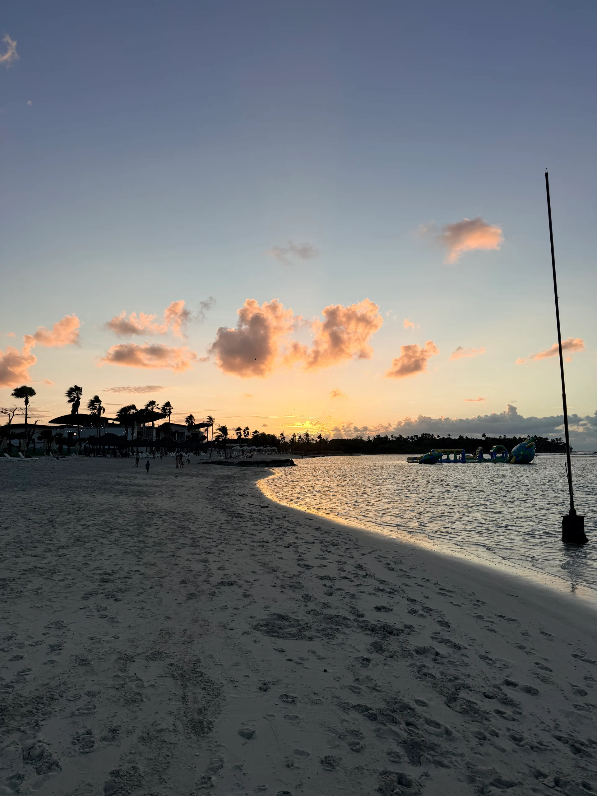 Une vue sereine du coucher de soleil sur une plage de sable blanc à Colón, au Panama. Le ciel est enflammé de teintes orange et rose, se reflétant sur l'eau calme. Des palmiers et quelques baigneurs ponctuent le paysage, créant une scène tropicale paisible.