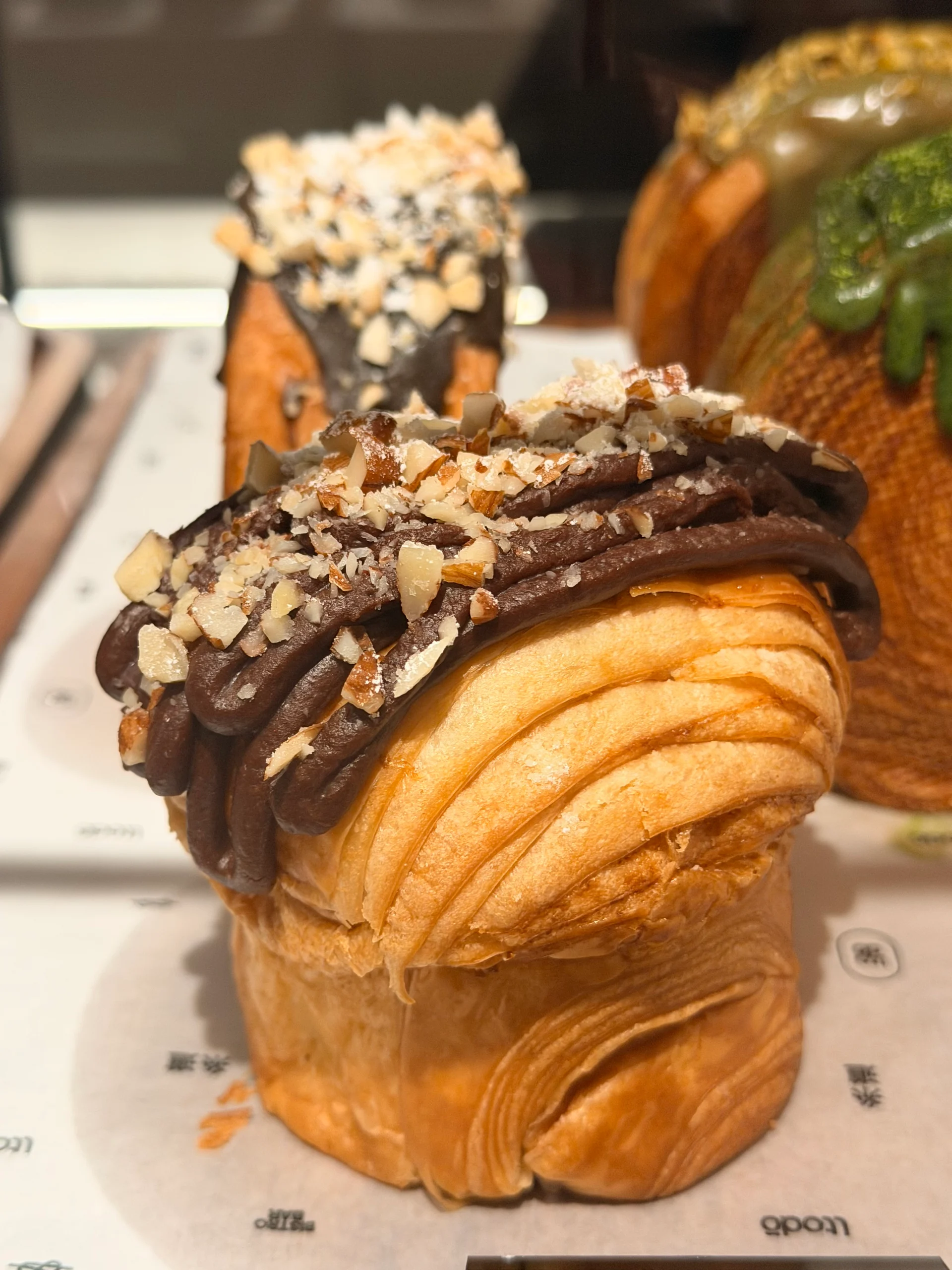 Close-up of a delectable chocolate croissant topped with a rich chocolate ganache and chopped nuts, displayed in a bakery window in Panama City.