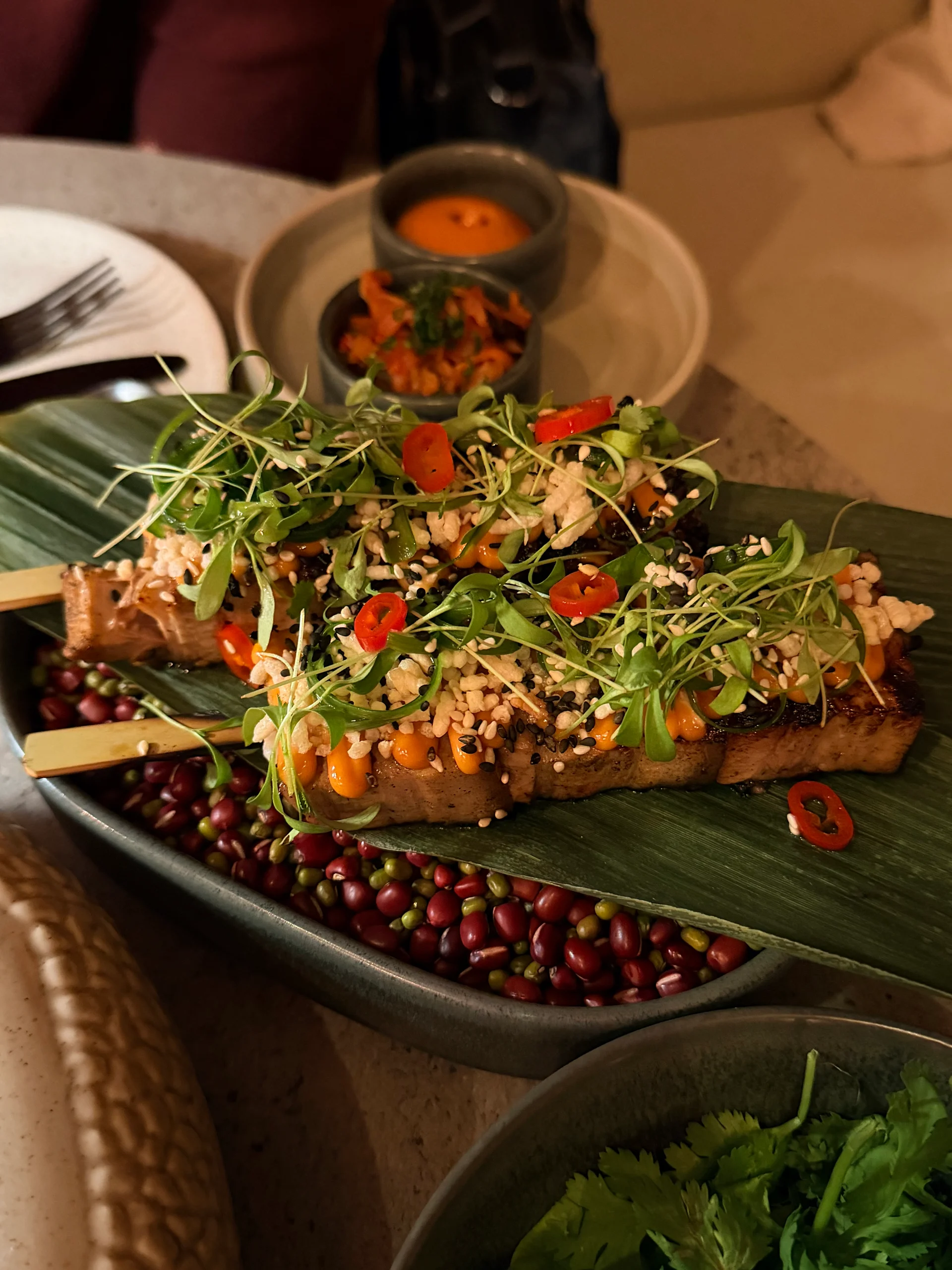 A beautifully presented grilled salmon dish garnished with microgreens, sesame seeds, and chili flakes, served on a bed of colorful beans and accompanied by a small bowl of vibrant sauce. The dish is presented on a large green leaf, adding a touch of nature to the upscale dining experience.