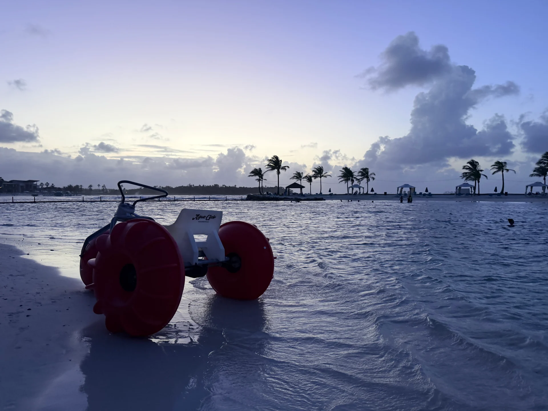 Un quad de plage rouge et blanc éclatant se trouve dans des eaux peu profondes au coucher du soleil sur Playa Escondida, Panama. Des palmiers et un complexe hôtelier sont visibles en arrière-plan sous un ciel dramatique rempli de nuages.