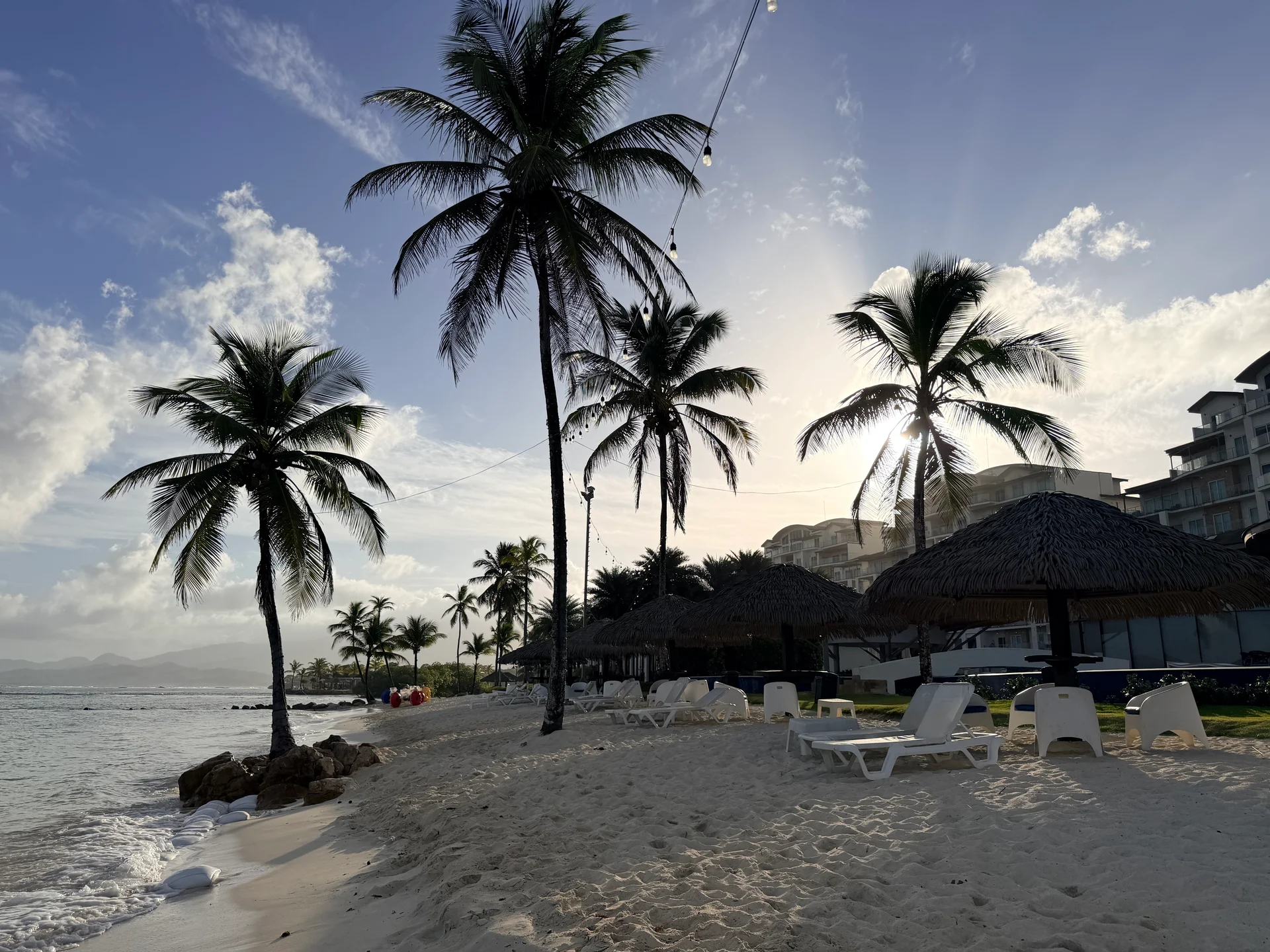 Une scène de plage sereine au coucher du soleil à Colón, au Panama. Des palmiers se balancent doucement le long de la plage de sable blanc, avec des chaises longues et des paillotes invitant à la détente. Un bâtiment de station balnéaire est visible en arrière-plan, complétant ce paradis tropical.