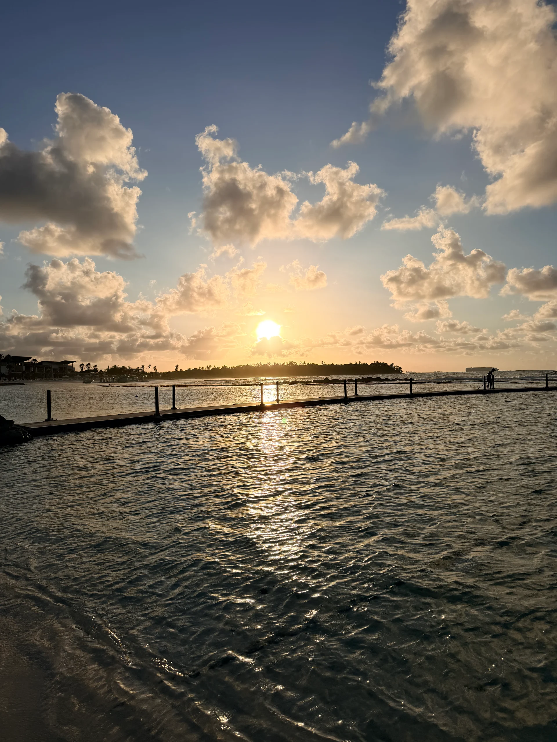 Une vue sereine du coucher de soleil sur une piscine calme et clôturée à Playa Escondida, Panama. La lumière dorée se reflète magnifiquement sur l'eau, créant une scène paisible et pittoresque. Des palmiers et des bâtiments lointains se profilent sur le ciel vibrant.