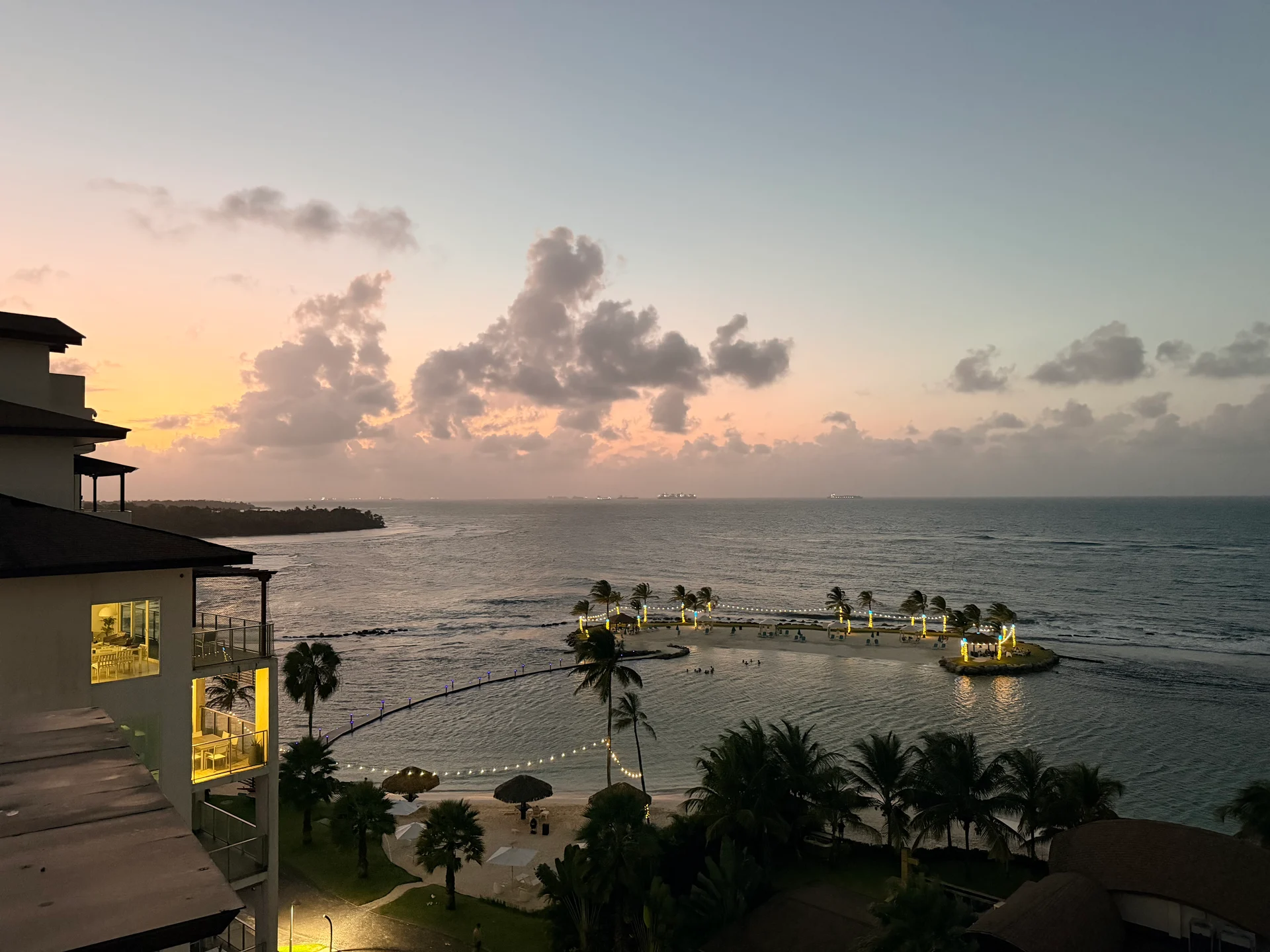 Une vue pittoresque du coucher de soleil sur l'océan tranquille à Playa Escondida Resort au Panama. La scène présente un bâtiment moderne avec un balcon surplombant l'eau, un charmant chemin orné de lumières menant à une petite île et de luxuriantes palmiers complétant le paradis tropical.