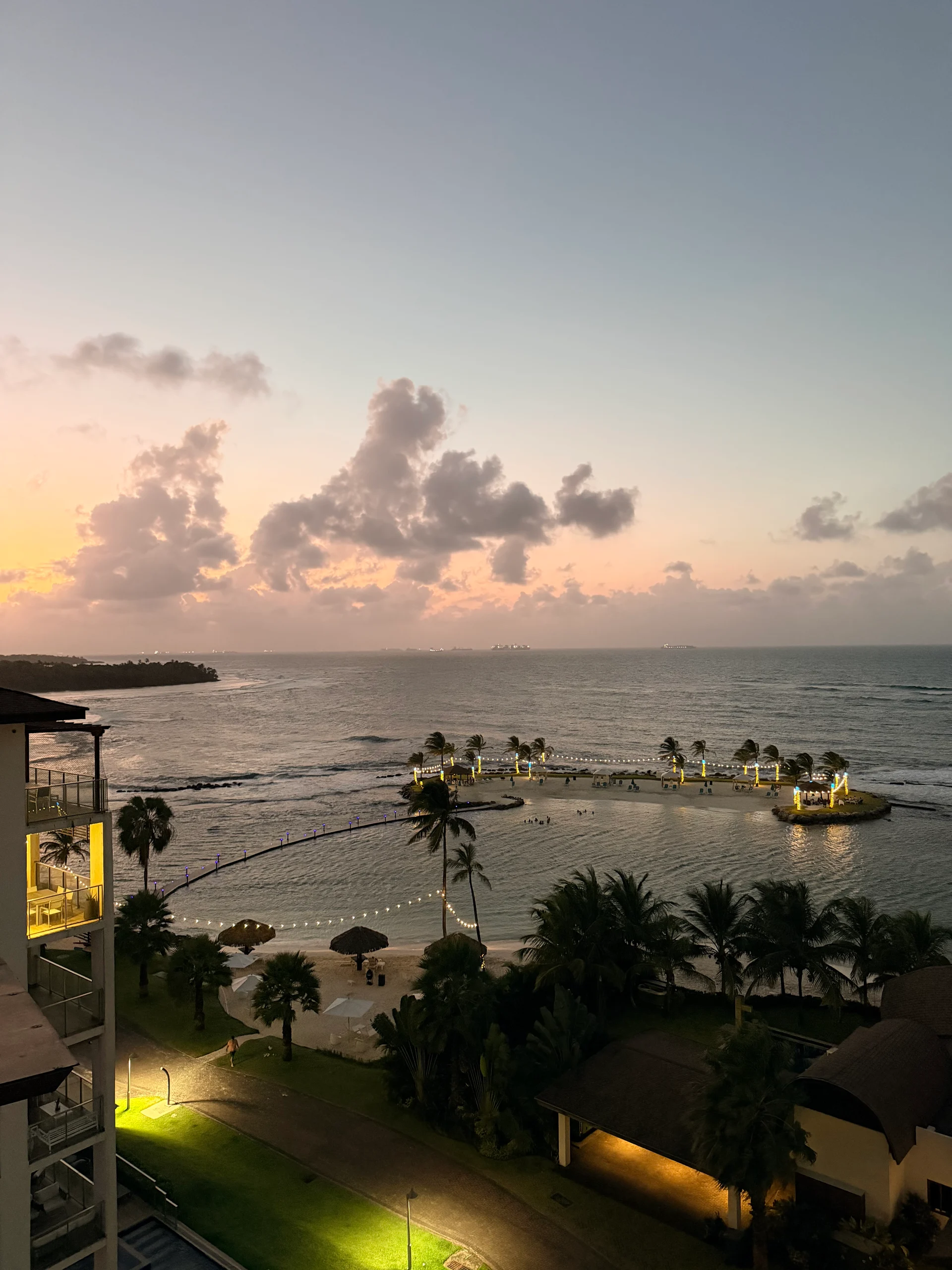 Une vue imprenable sur le coucher de soleil au-dessus du Resort Playa Escondida, Panama. Des palmiers et une végétation luxuriante encadrent une scène de plage paisible avec des vagues douces et des guirlandes lumineuses illuminant le rivage. Le ciel est embrasé de chaudes teintes d'orange et de rose, créant une atmosphère sereine et invitante.