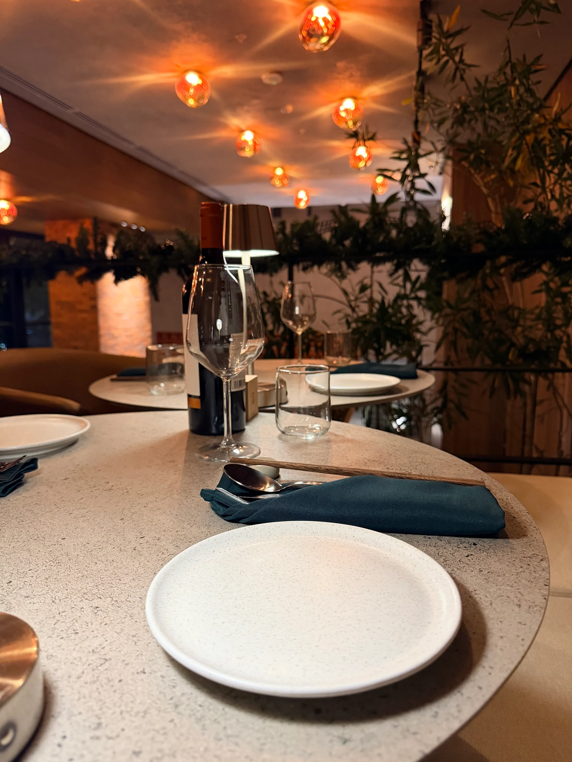 Elegant restaurant setting featuring a table set with glassware, a wine bottle, a plate, and silverware. Warm lighting and decorative foliage create a sophisticated atmosphere.