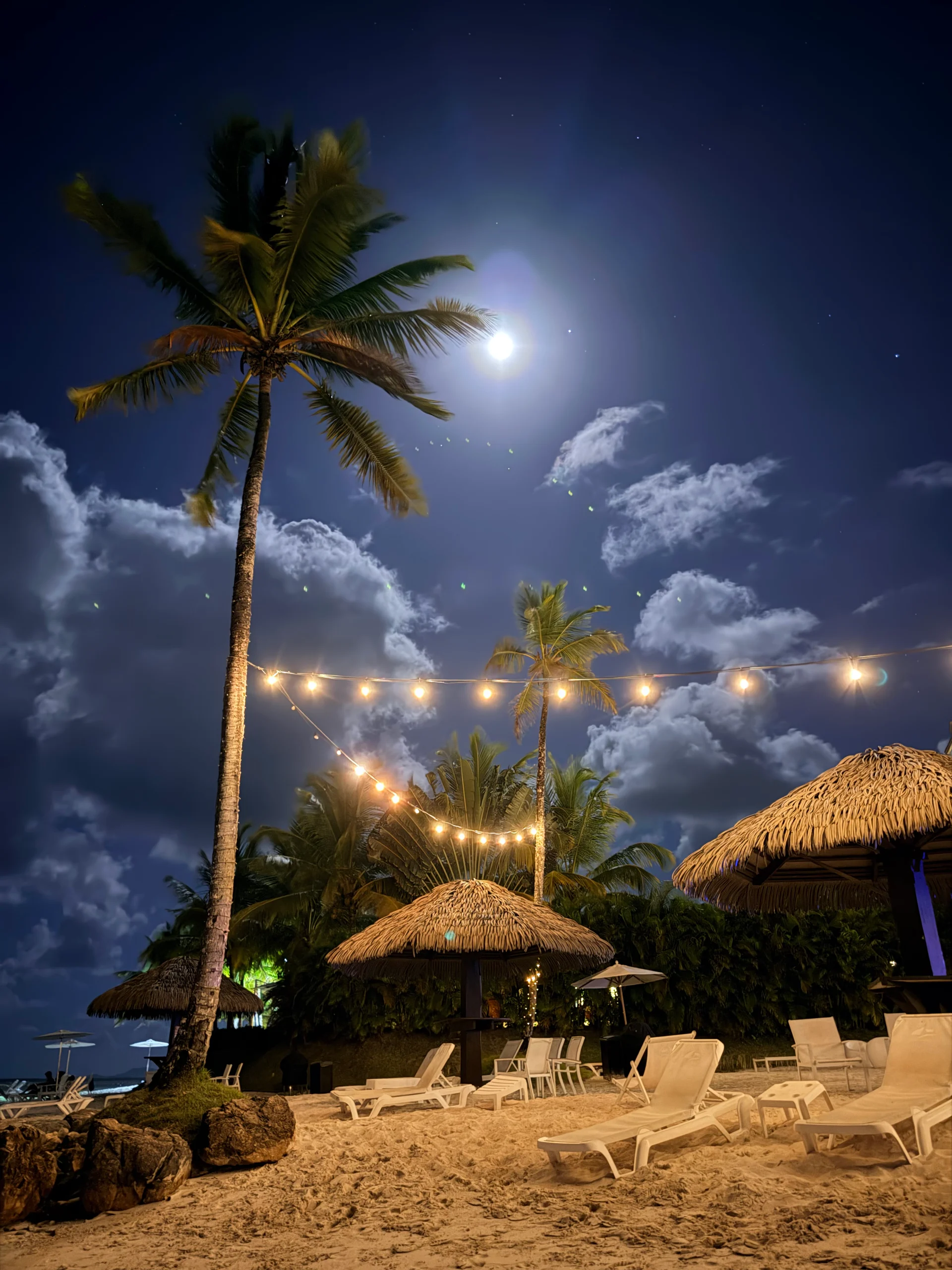 Une scène nocturne sereine à Playa Escondida, au Panama, avec des palmiers se détachant sur une lune brillante et des guirlandes lumineuses illuminant des paillotes et des chaises longues sur une plage de sable. L'atmosphère paisible évoque un sentiment de paradis des Caraïbes.
