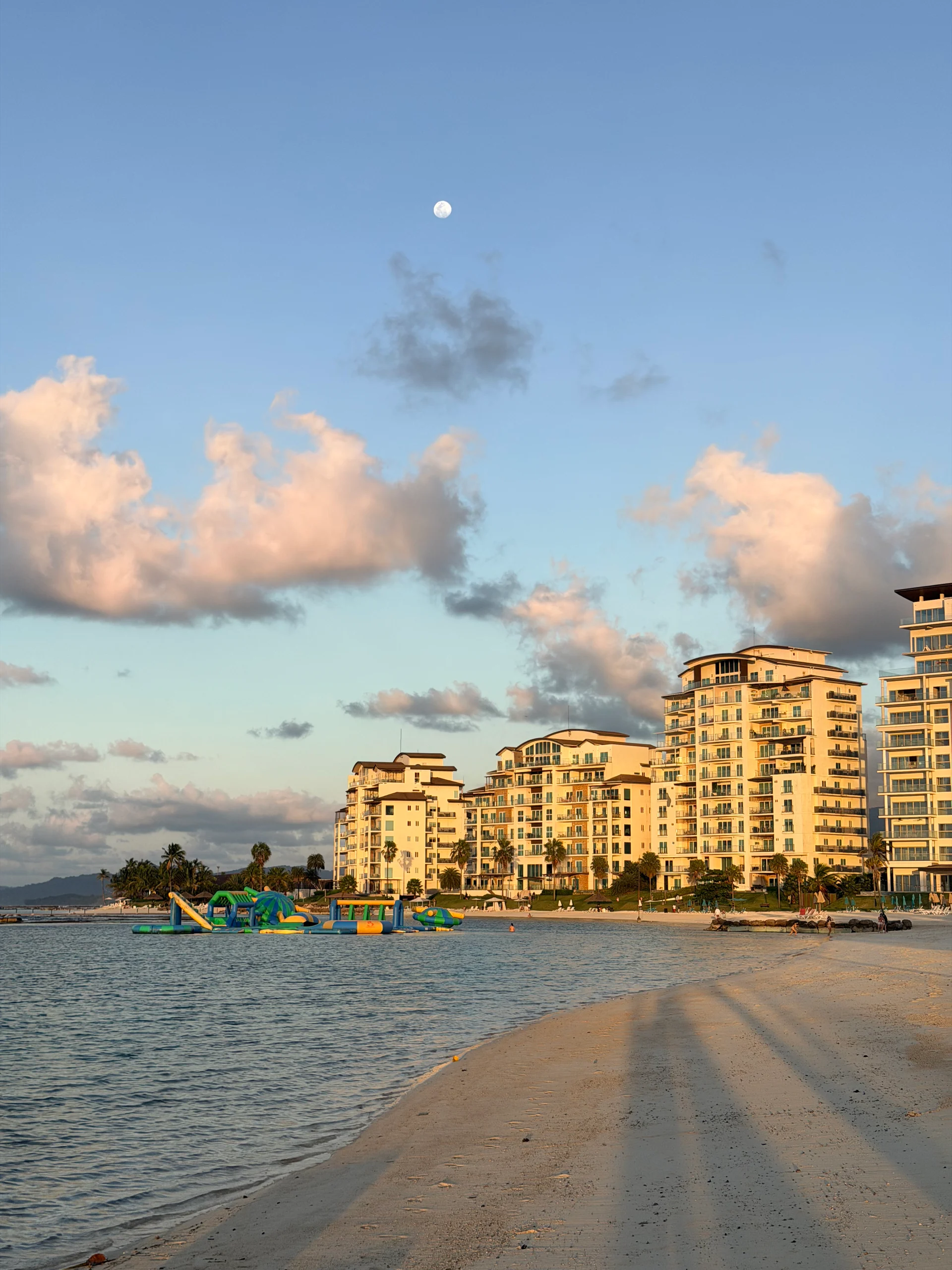 Une vue côtière sereine à Playa Escondida, au Panama, mettant en valeur un vaste complexe hôtelier baigné de la douce lumière du crépuscule. Les eaux turquoise calmes lèchent doucement une plage de sable blanc immaculée, reflétant les chaudes teintes du coucher de soleil et une lune pleine lumineuse. Un parc aquatique gonflable vibrant ajoute une touche ludique à la scène.