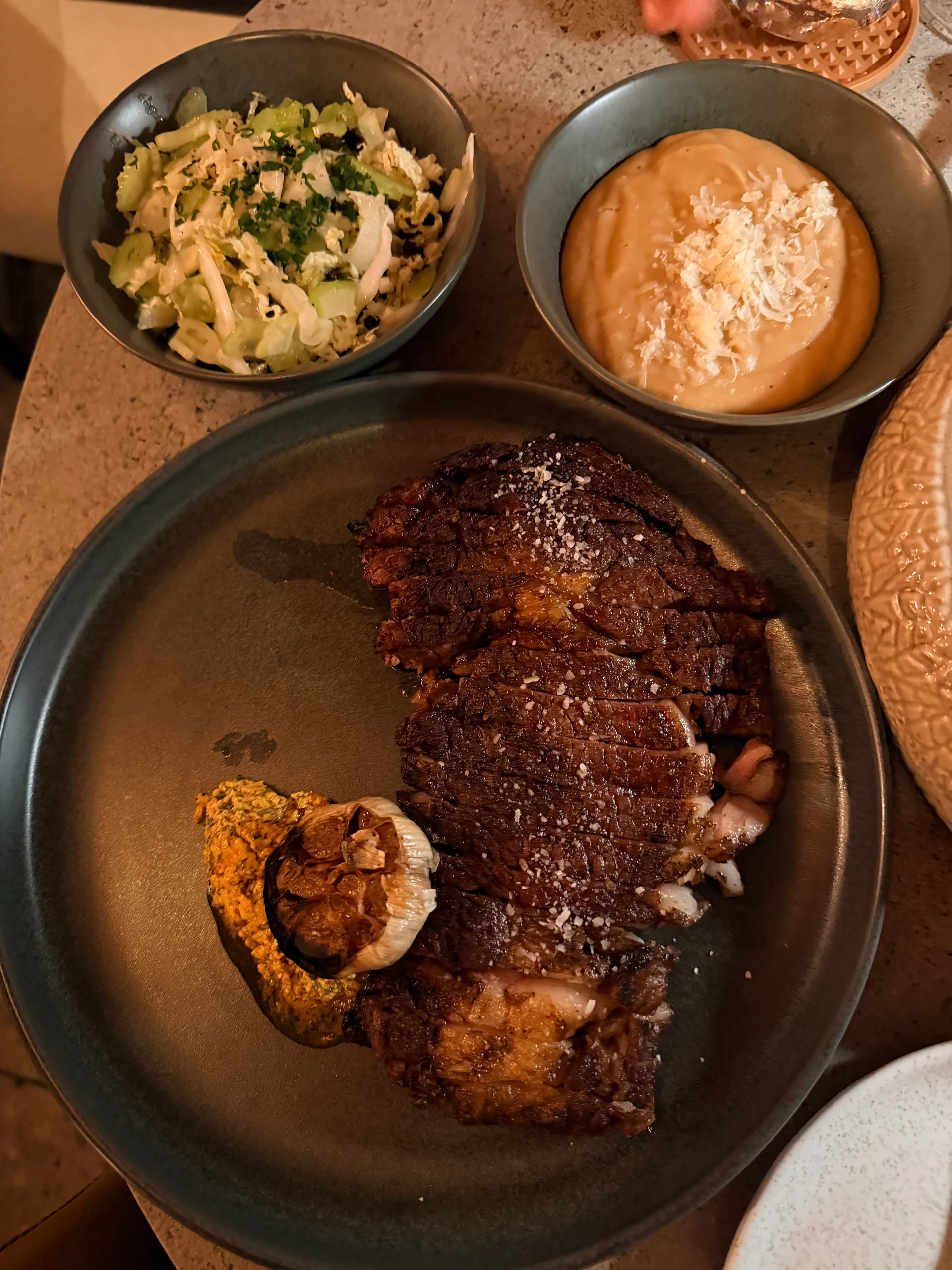 A beautifully plated steak dinner at Dovle Cincuentenario restaurant in Panama City, featuring a perfectly grilled steak with roasted garlic, accompanied by a vibrant slaw and creamy purée. The warm lighting creates an inviting and upscale dining experience.