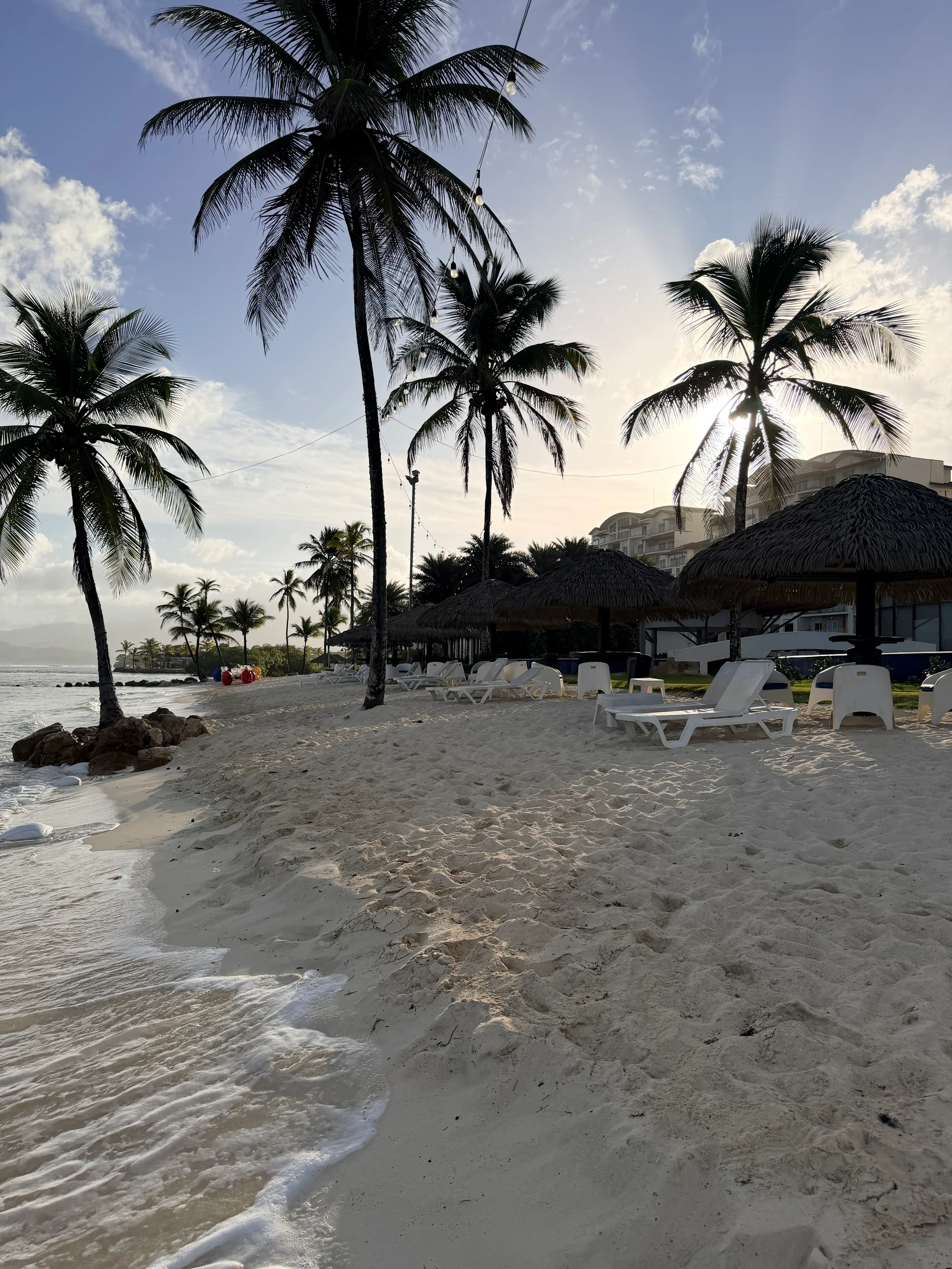 Une scène de plage sereine à Playa Escondida, au Panama, avec des palmiers, du sable blanc et des cabanas au toit de chaume avec des chaises longues. Le soleil se couche, projetant une lueur chaude sur les eaux tranquilles.