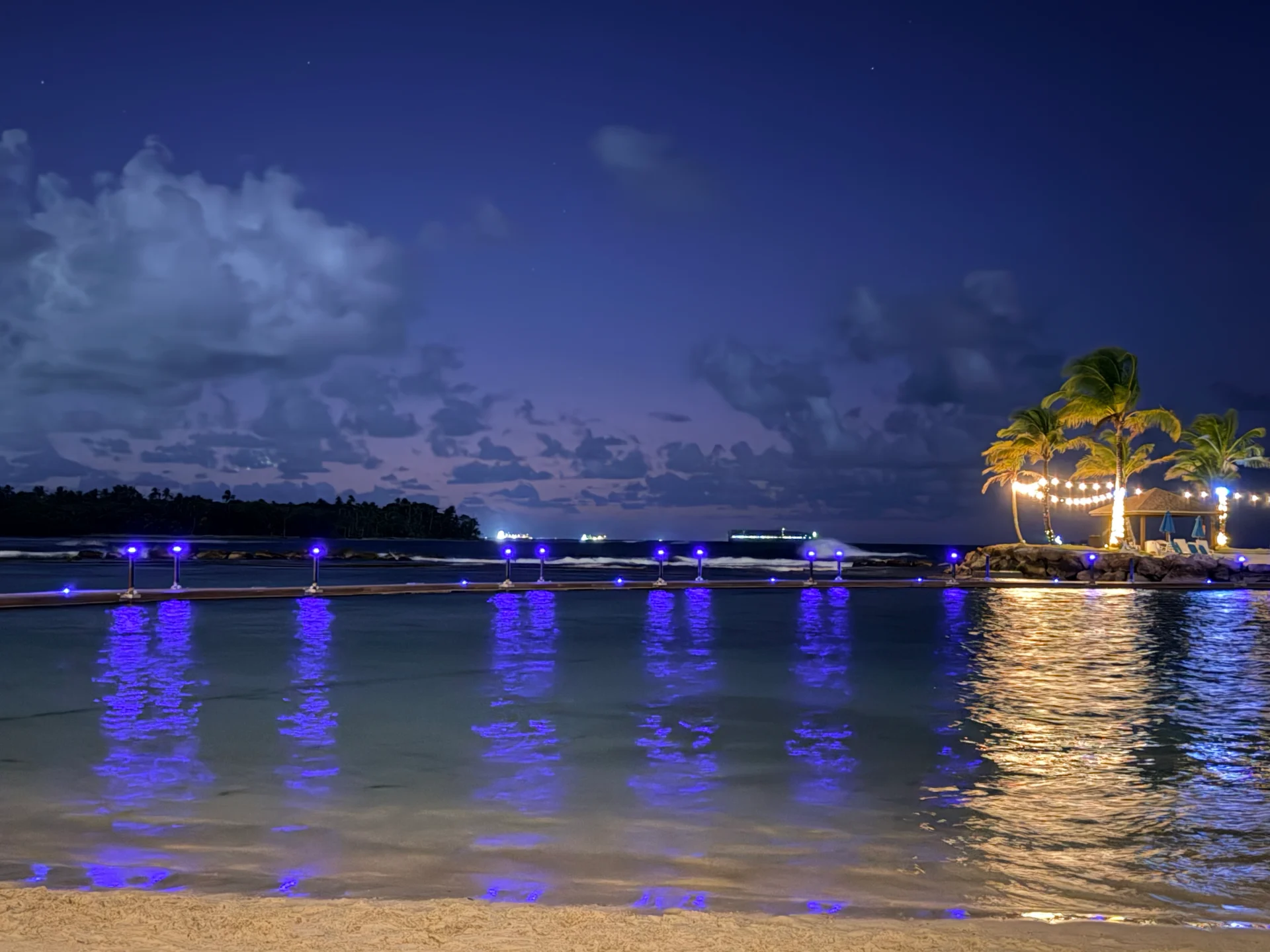 Scène nocturne paisible à Playa Escondida, Panama. Des lumières bleues douces illuminent les eaux calmes, se reflétant dans le ciel et le sable. Un palmier et des guirlandes lumineuses ajoutent à l'atmosphère sereine et tropicale.