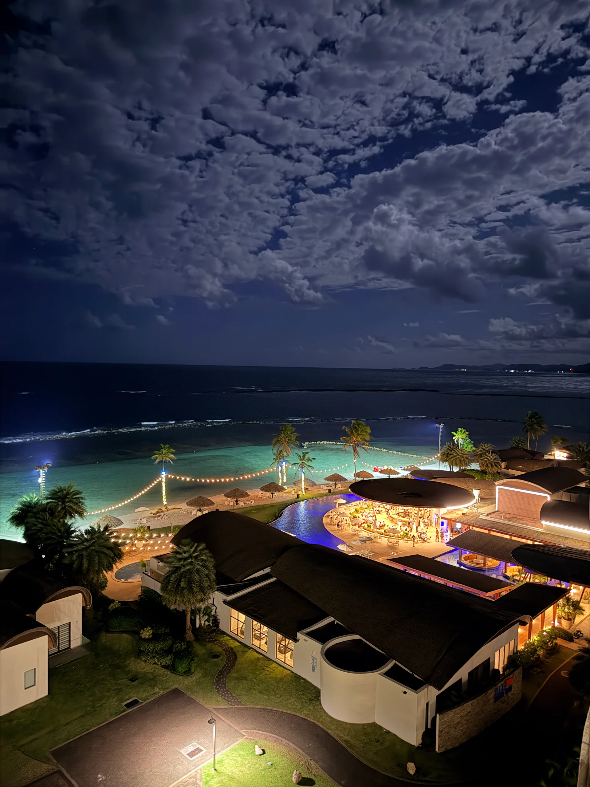 Vue aérienne nocturne du Resort & Marina Playa Escondida au Panama, présentant une zone de piscine éclairée, une plage avec des guirlandes lumineuses et une architecture moderne contre un ciel nuageux spectaculaire.