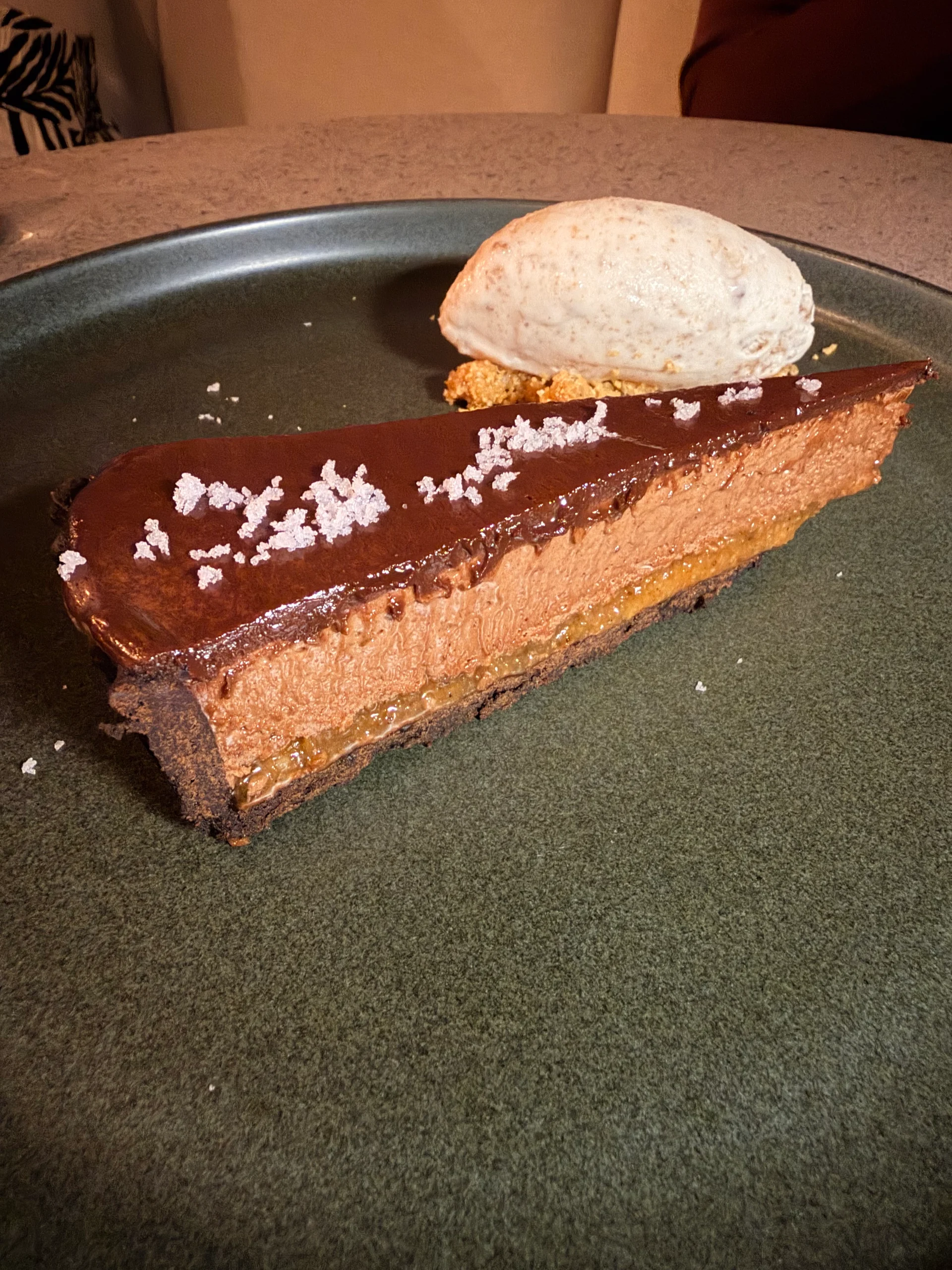 A close-up, mouthwatering slice of layered chocolate caramel cake topped with flaky sea salt and a scoop of creamy vanilla ice cream, presented on a dark green plate.