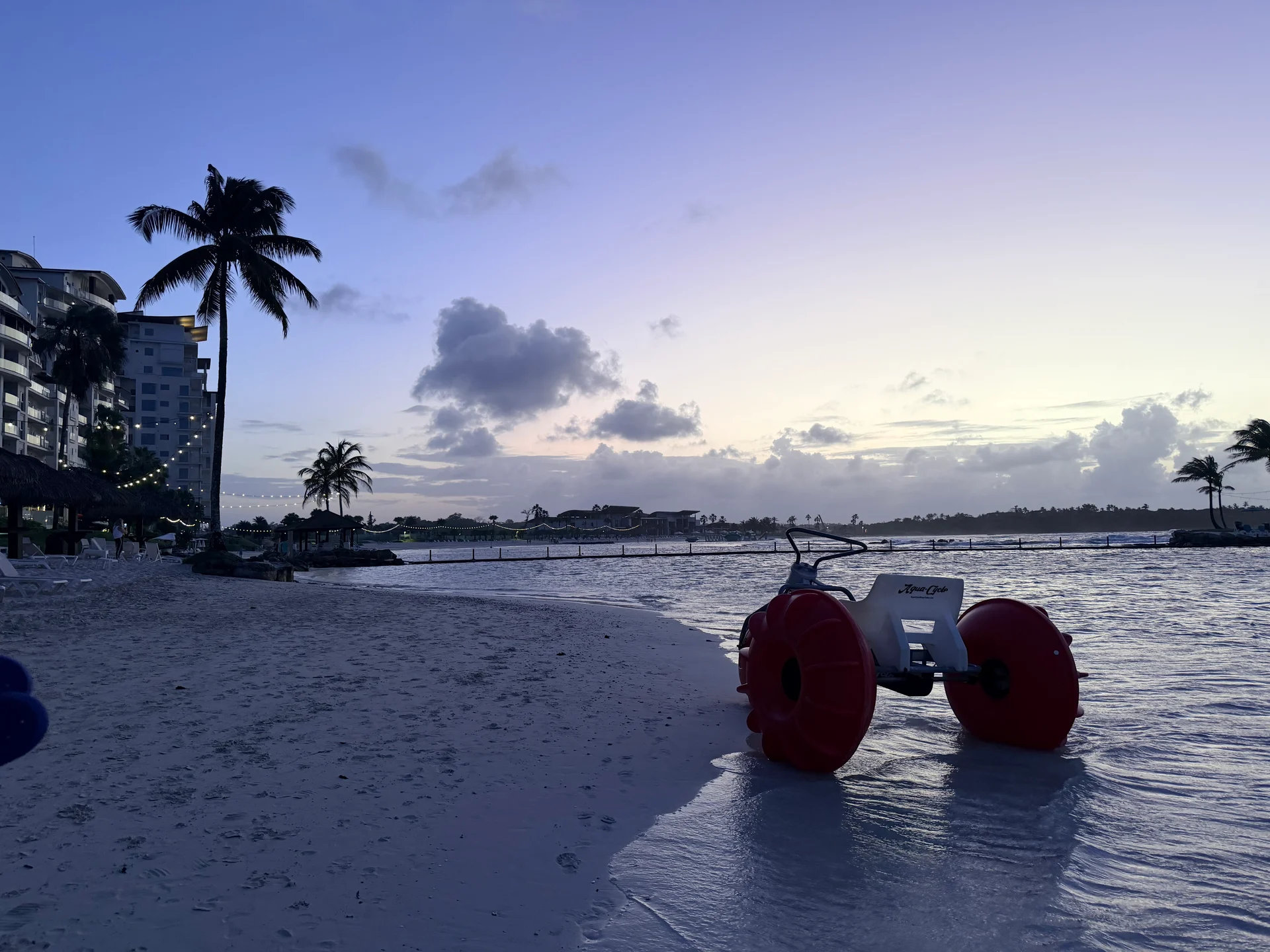 Une scène sereine au crépuscule sur la plage de Playa Escondida, à Panama. Un véhicule amphibie rouge et blanc se trouve sur le sable mouillé, reflétant les couleurs du coucher de soleil. L'architecture caribéenne moderne et les palmiers sont visibles en arrière-plan, créant une atmosphère paisible et pittoresque.